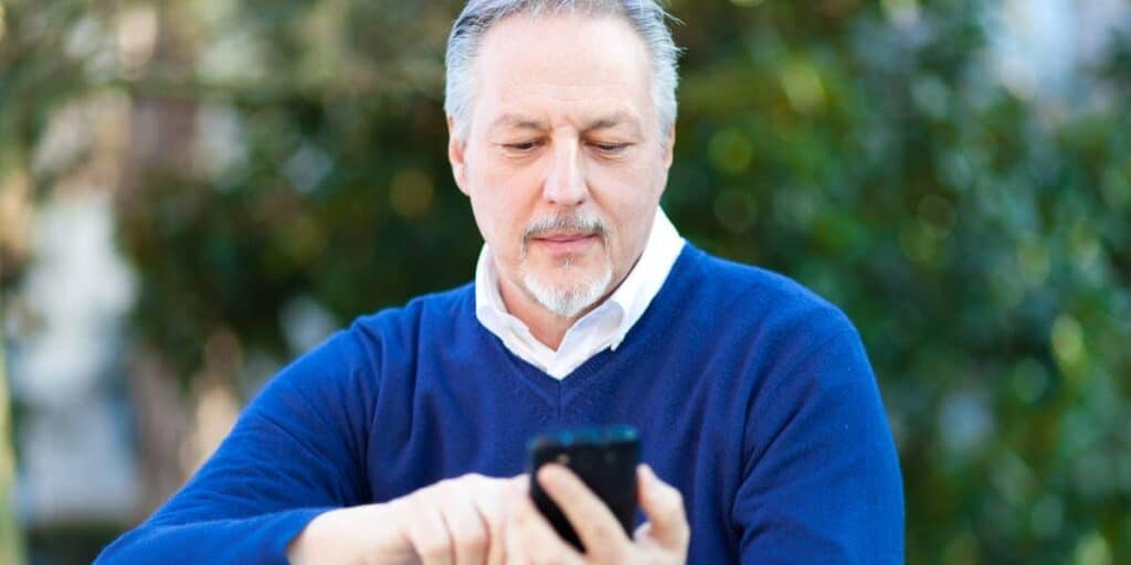 A senior man with gray hair and beard using smartphone outdoors for eye health awareness.