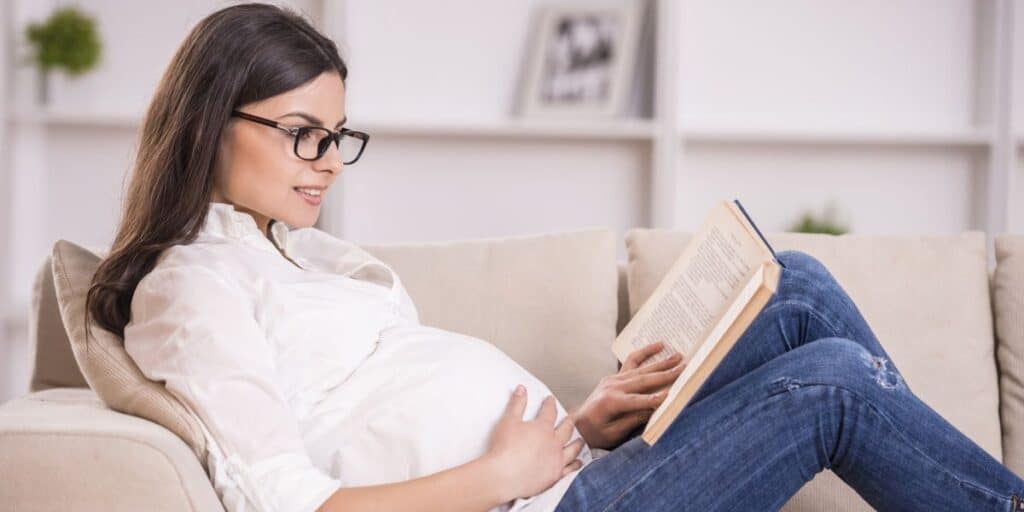 Pregnant woman enjoying reading comfortably at home with glasses.