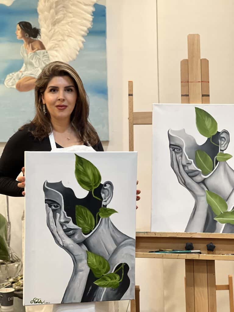 Woman holding a painting of a woman's face with green leaves, art studio setting.